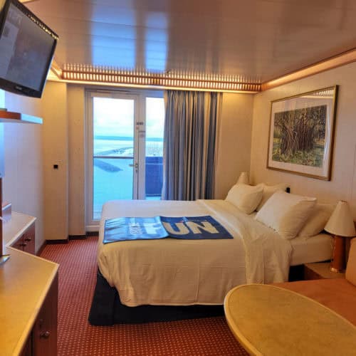 Carnival stateroom with picture window and balcony, bed with choose fun mat on top of it, and television on the wall