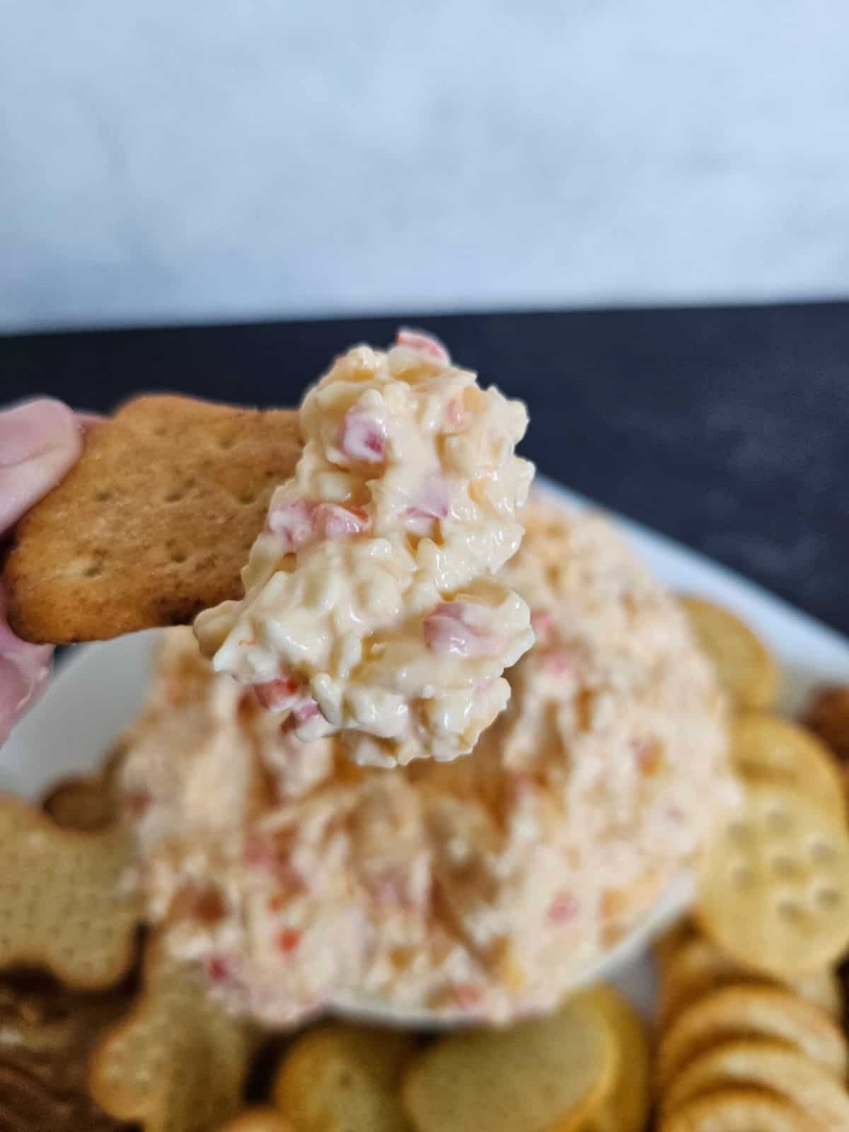 Pimento cheese on a cracker being held above a bowl of cheese and plate of crackers