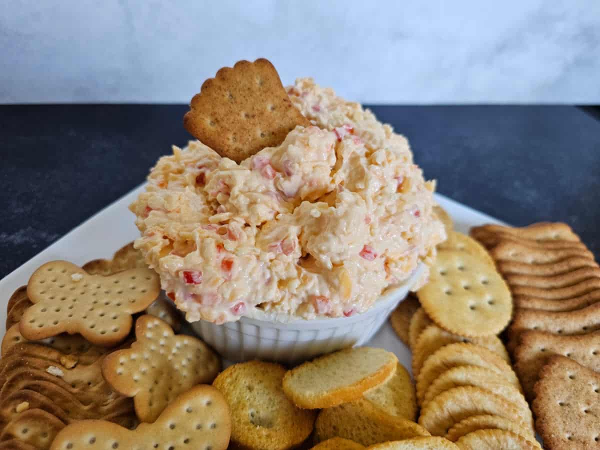 wheat cracker in a bowl of pimento cheese on a white plate filled with a variety of crackers